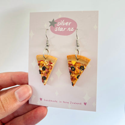 A hand presents a card displaying Pizza Polymer Clay Earrings, crafted with pizza slice designs complete with toppings such as olives and peppers. The card is labeled "silverstar.nz" and notes "handmade in New Zealand," featuring stainless steel hooks along with decorative stars and hearts.