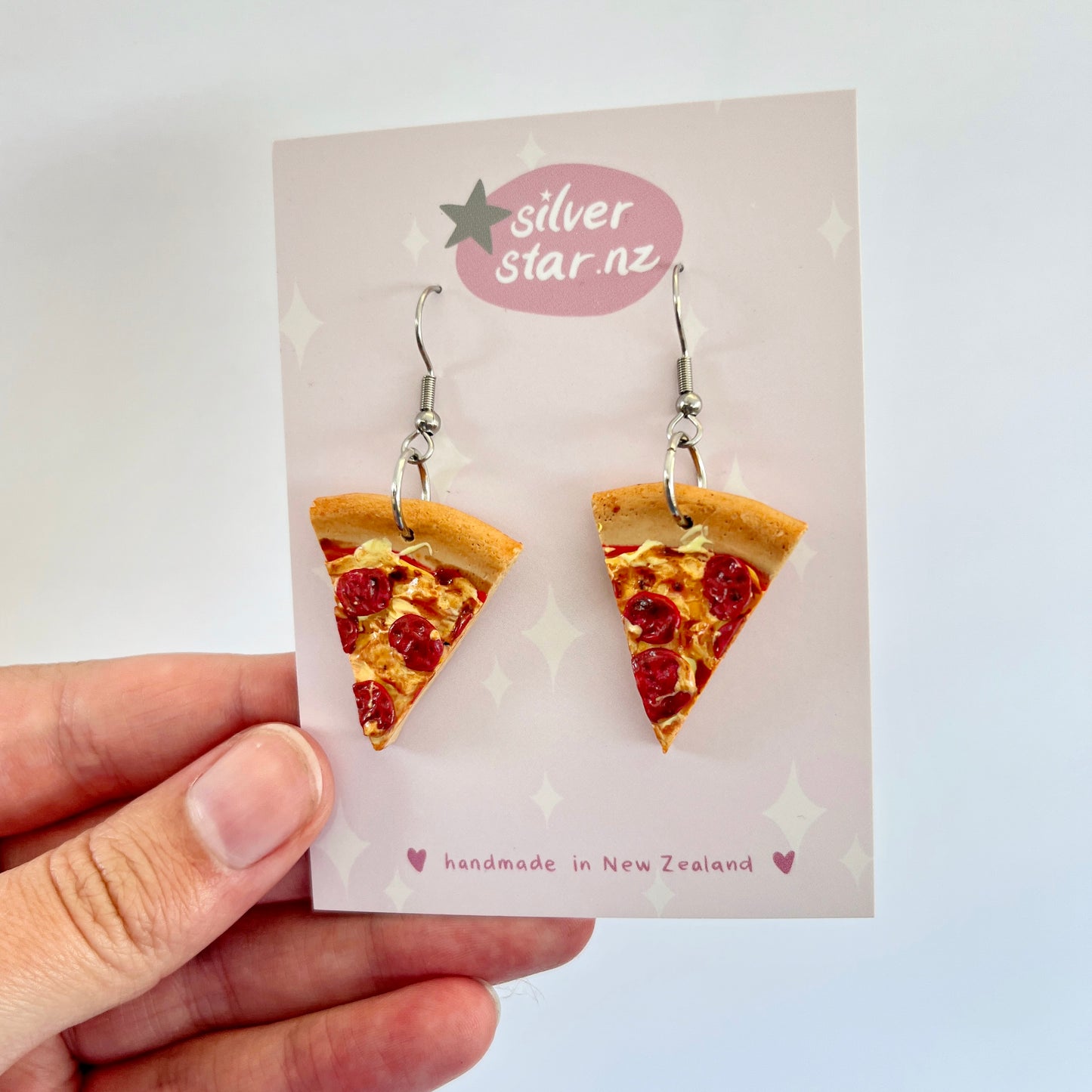 A hand displays a card featuring the Pizza Polymer Clay Earrings, crafted by silverstar.nz. These handmade earrings, shaped like pepperoni pizza slices, boast stainless steel hooks and are thoughtfully made in New Zealand.