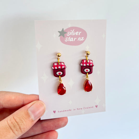 A hand holds a card displaying the Berry Jam Jar Polymer Clay Earrings, carefully crafted with intricate designs. The earrings feature charming red telephone booth motifs, enhanced by teardrop-shaped red gems hanging from stainless steel hooks. The card prominently features the silverstar.nz logo and proudly highlights their creation in New Zealand.