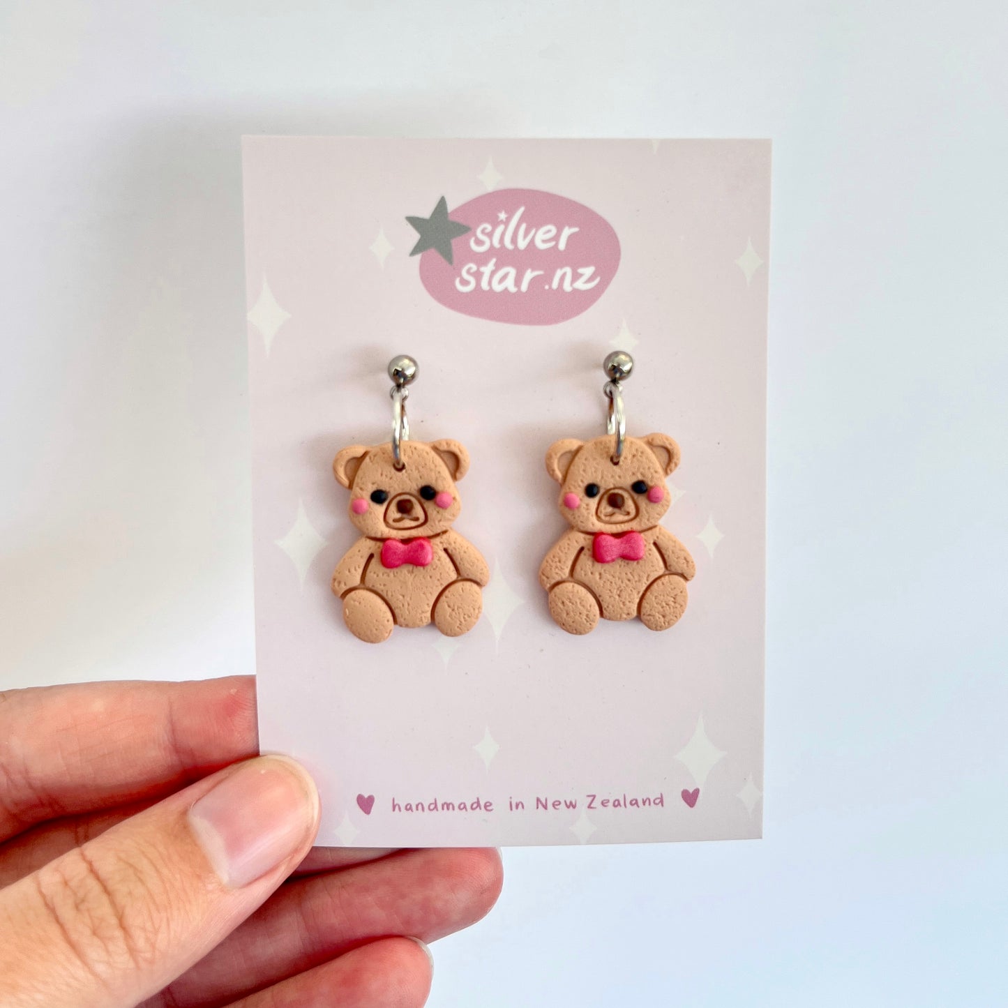 A close-up shows a hand holding a card featuring the funky design of the Teddy Polymer Clay Earrings, shaped like teddy bears. These light brown bears, adorned with pink bows, dangle from stainless steel hooks. The card displays "silverstar.nz" and notes they are handmade in New Zealand.