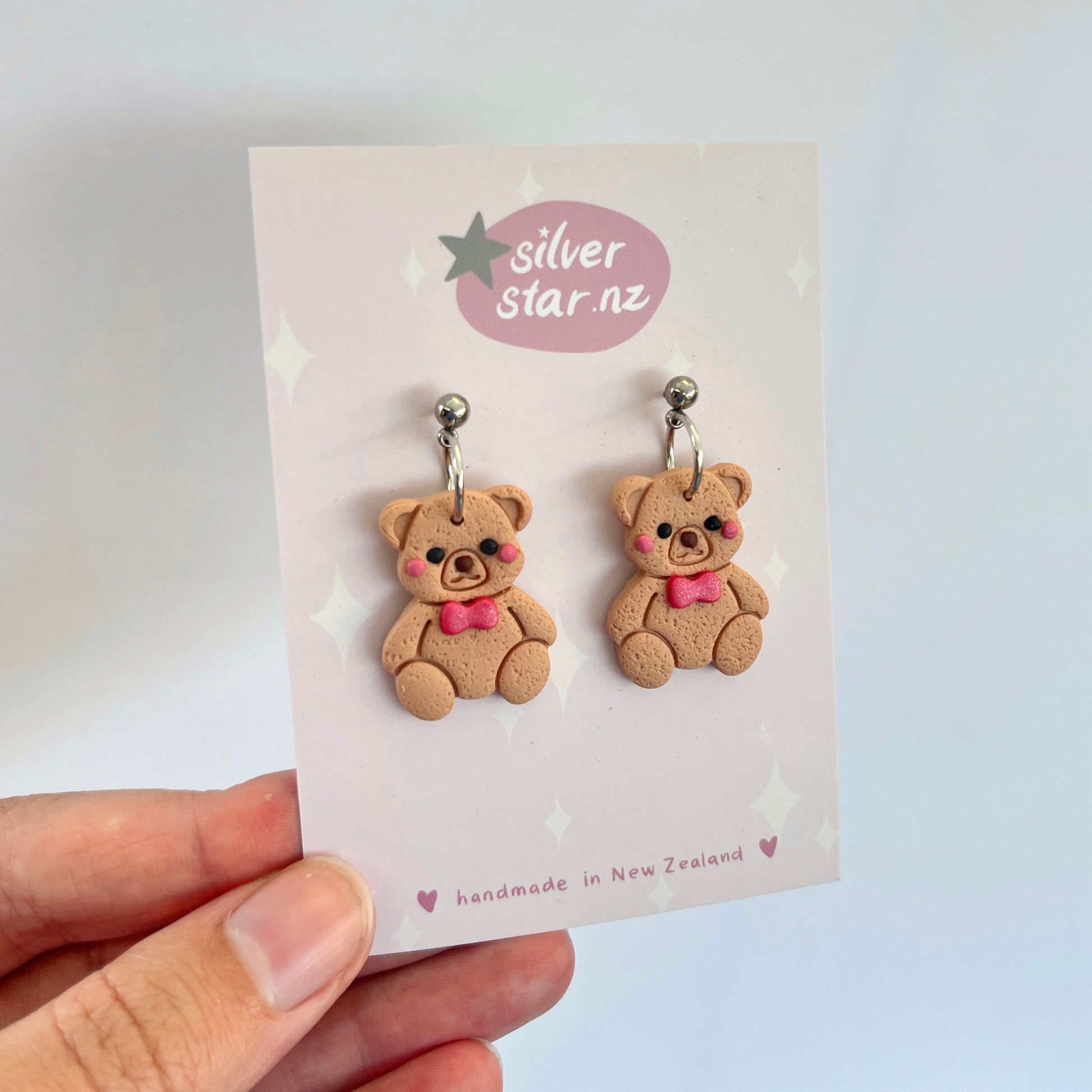 A hand holds up a card featuring teddy bear earrings with pink bows, crafted from handmade polymer clay. The card displays "silverstar.nz" and emphasizes the unique, funky design and stainless steel hooks, all meticulously crafted in New Zealand.