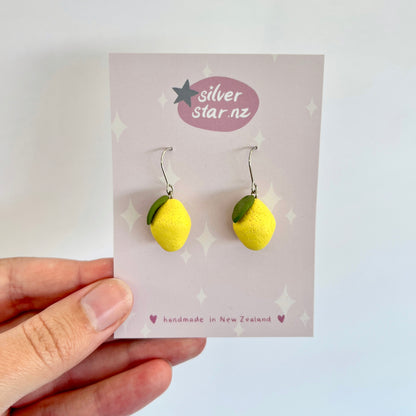 A person's hand holds a card displaying the Mini Lemon Polymer Clay Earrings, which feature unique, handmade designs of lemons with green leaf accents. The card is branded silverstar.nz and marked as handmade in New Zealand, showcasing the charming craftsmanship of these delightful accessories.