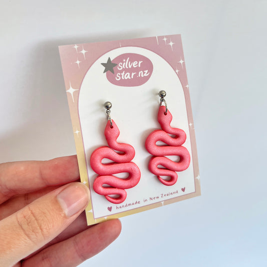 A hand displays the Snake Polymer Clay Earrings from silverstar.nz, featuring pink, snake-shaped designs. These earrings are crafted with polymer clay and come with durable stainless steel hooks. They are presented on a pastel pink card adorned with decorative stars and labeled "handmade in New Zealand.