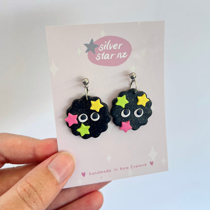 A hand showcases a pair of Soot Sprite Polymer Clay Earrings, featuring googly eyes and colorful star decorations, crafted by silverstar.nz. These unique pieces are handmade in New Zealand and displayed on a card reflecting their brand origin.