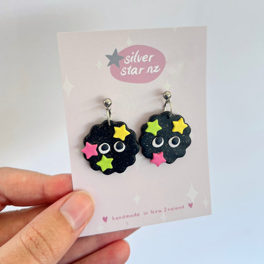 A hand showcases a pair of Soot Sprite Polymer Clay Earrings, featuring googly eyes and colorful star decorations, crafted by silverstar.nz. These unique pieces are handmade in New Zealand and displayed on a card reflecting their brand origin.