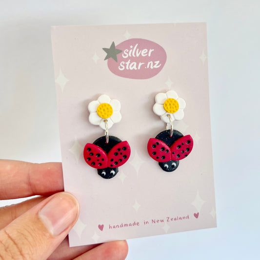 A hand holds a card displaying the Ladybird Polymer Clay Earrings by silverstar.nz. The design includes a daisy with white petals and a yellow center, complemented by a red ladybug with black spots. The earrings, which are made in New Zealand, feature stainless steel hooks.