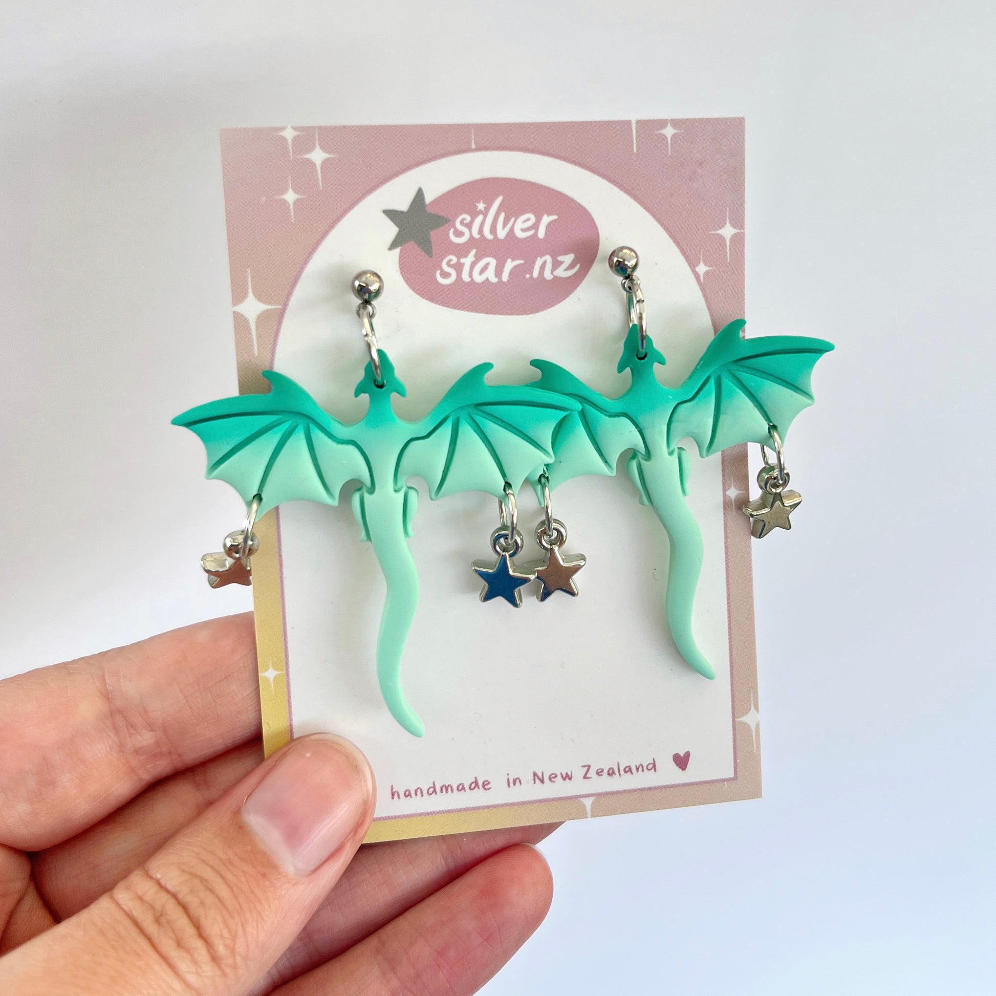 A hand holds a card displaying the striking (Imperfect) Mint Dragon Polymer Clay Earrings. These turquoise beauties are adorned with dangling star charms and are lovingly crafted in New Zealand. The card features the brand name silverstar.nz, highlighting the expertly handmade nature with a heart icon.