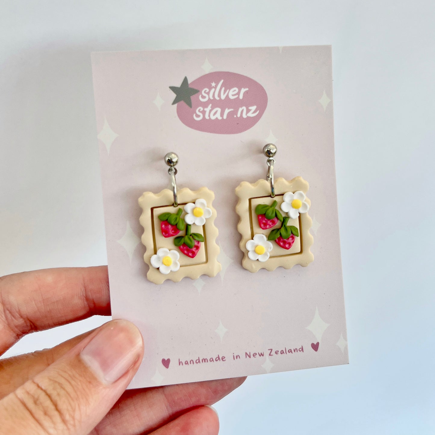 A hand presents the Strawberry Stamp Polymer Clay Earrings, featuring a charming square biscuit design embellished with strawberries and white flowers. Crafted by silverstar.nz in New Zealand, these handmade earrings come with stainless steel hooks for durability.