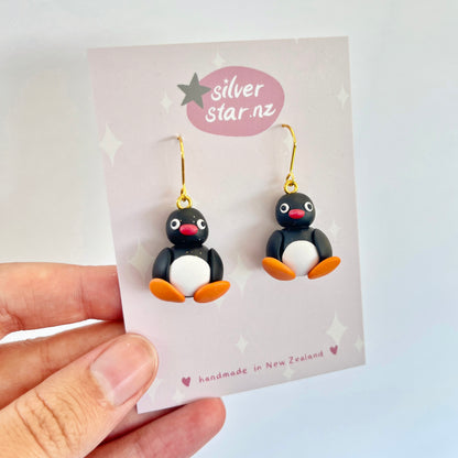 A hand holds a card showcasing the Pingu Polymer Clay Earrings, which are uniquely handmade into the shape of penguins with black bodies, white bellies, and orange beaks and feet. The card features the brand silverstar.nz and confirms they are handmade in New Zealand.