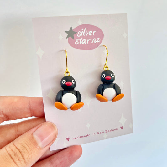 A hand holds a card showcasing the Pingu Polymer Clay Earrings, which are uniquely handmade into the shape of penguins with black bodies, white bellies, and orange beaks and feet. The card features the brand silverstar.nz and confirms they are handmade in New Zealand.