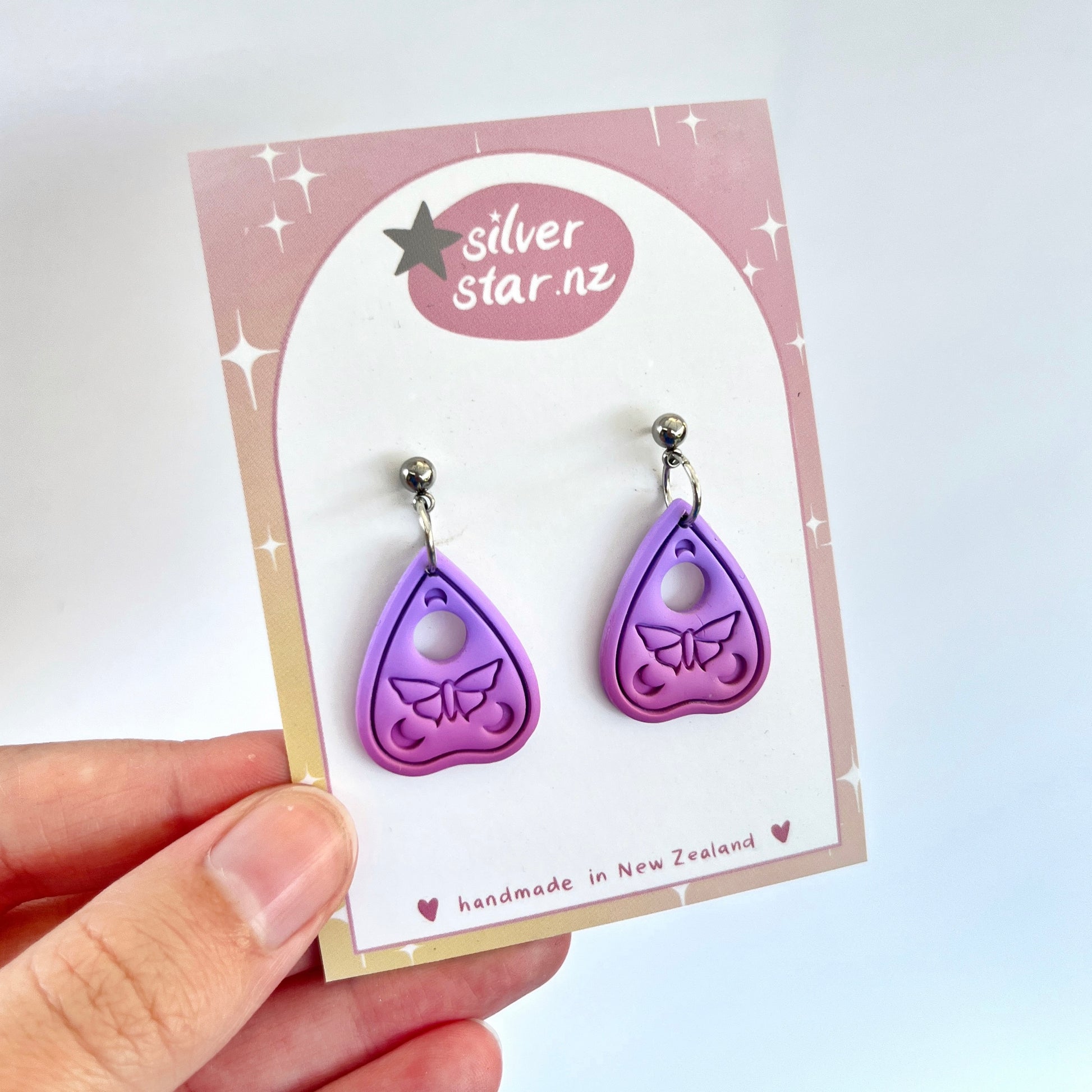 A hand holds a card showcasing a distinctive pair of Ouija Planchette Polymer Clay Earrings, intricately designed with butterfly cutouts in a unique purple teardrop shape. The card highlights the brand silverstar.nz and indicates that these handcrafted earrings are made in New Zealand.