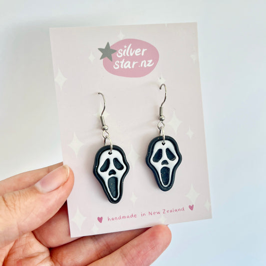 A hand holds a pair of Ghostface Polymer Clay Earrings, featuring stainless steel hooks, displayed on a card labeled silverstar.nz. These New Zealand handmade earrings are crafted from black and white polymer clay and are surrounded by small star and sparkle designs.