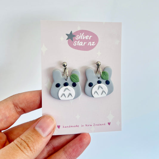 Close-up of a hand holding funky Totoro-shaped earrings on a silverstar.nz branded card. These handmade Totoro earrings feature gray faces with green details. The card showcases a purple logo and text declaring they are crafted from polymer clay in New Zealand.