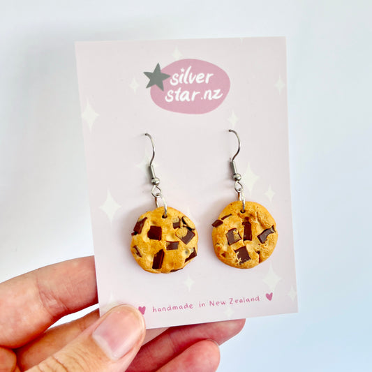 A hand presents a card displaying the Chocolate Chip Cookie Earrings, skillfully made from polymer clay to look like chocolate chip cookies. The card features a star emblem and the text "silverstar.nz," highlighting that these handmade earrings originate from New Zealand.