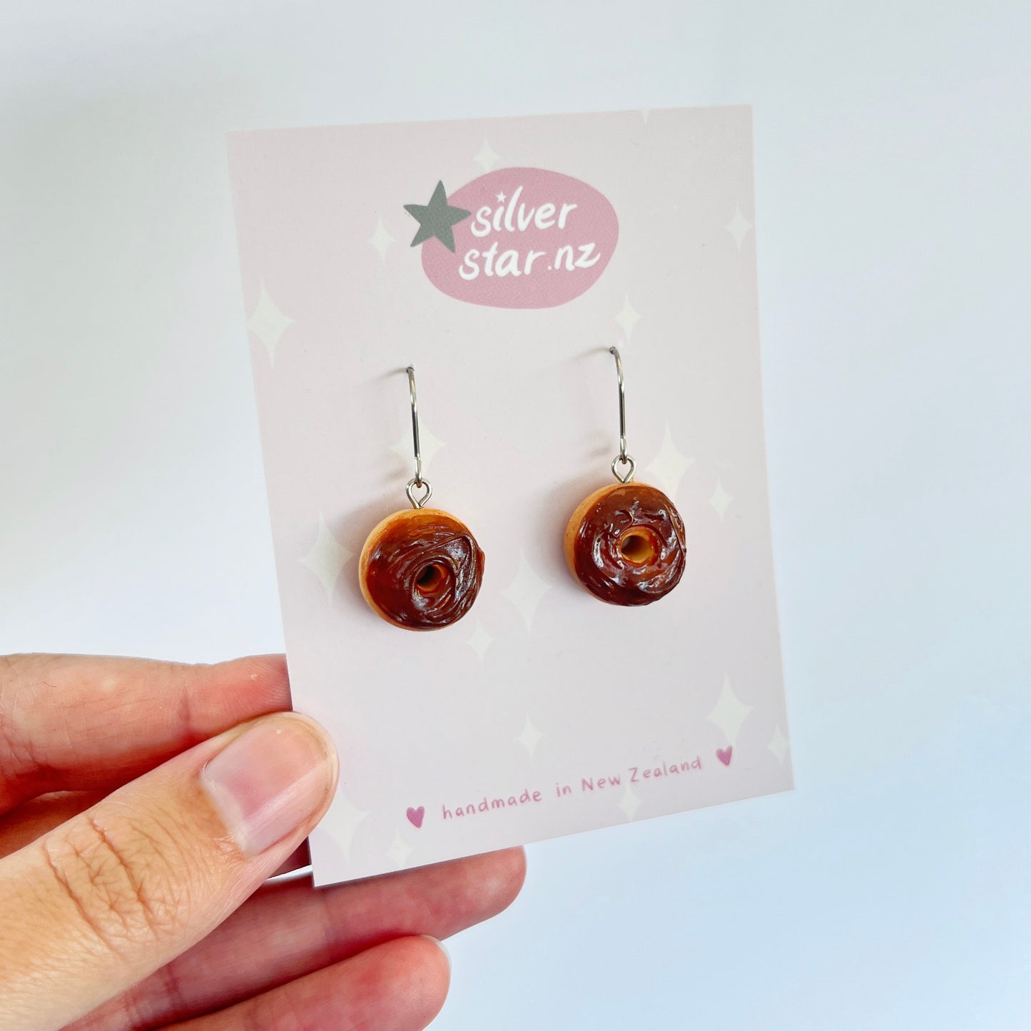Close-up of a hand holding a card with unique polymer clay earrings shaped like chocolate iced donuts. The card features a silverstar.nz logo and the text "handmade in New Zealand." These handmade earrings have hooks for pierced ears.