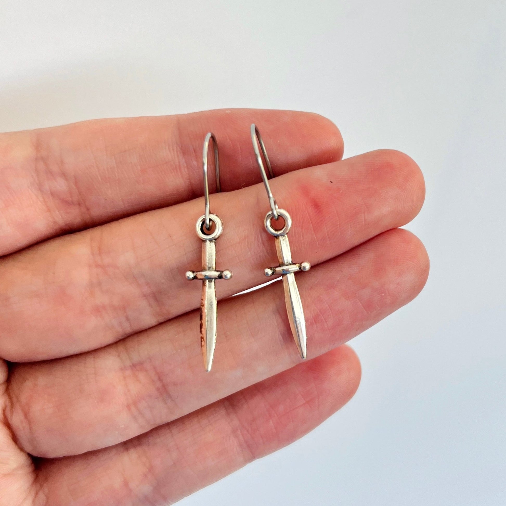 A hand presents a pair of Mini Silver Dagger Earrings by silverstar.nz on a white background. These lightweight earrings, designed to resemble miniature swords, feature intricate hilt details and are equipped with stainless steel hooks for added comfort and durability.