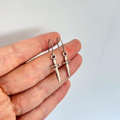 A hand showcases the Mini Silver Dagger Earrings by silverstar.nz, featuring a pair of dagger-shaped earrings with stainless steel hooks against a plain white background. These distinctive, hand-assembled earrings are notable for their unique design and meticulous craftsmanship.