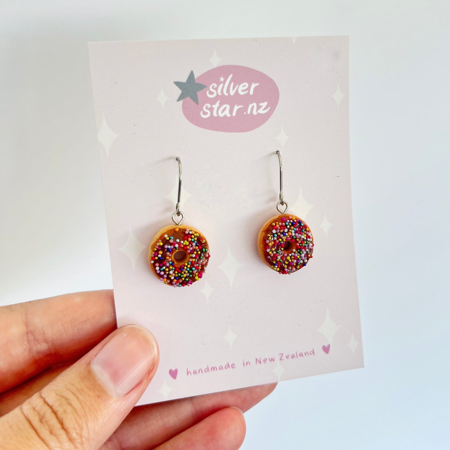 A hand presents a card featuring the Choc Iced Donut Polymer Clay Earrings, distinguished by their donut shape adorned with colorful sprinkles. The card, set against a star-patterned background, reads "silverstar.nz" and "handmade in New Zealand.