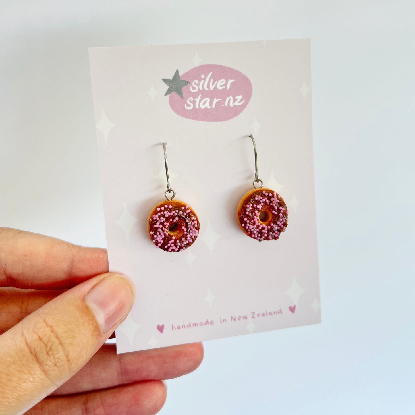 The Choc Iced Donut Polymer Clay Earrings, featuring a unique donut shape with brown frosting and pink sprinkles, are displayed against a white card that bears the brand name silverstar.nz. This card, decorated with a small gray star, emphasizes that these handmade earrings are crafted in New Zealand.