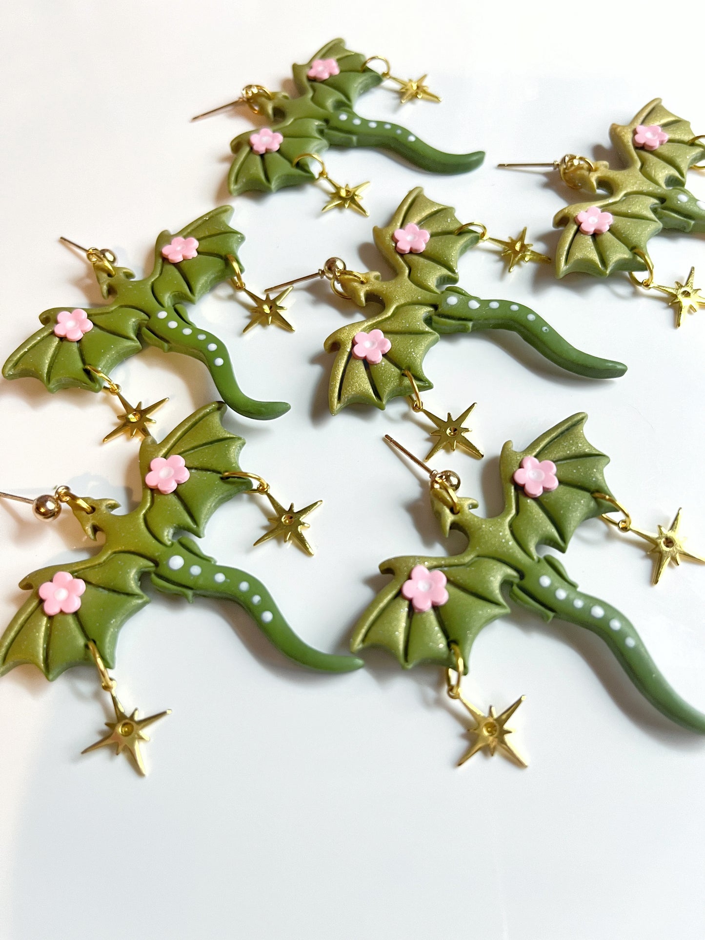 Close-up of silverstar.nzs Green Fae Dragon Polymer Clay Earrings featuring detailed wings, dotted tails, pink flower accents, and dangling gold stars on a white surface for a whimsical design.