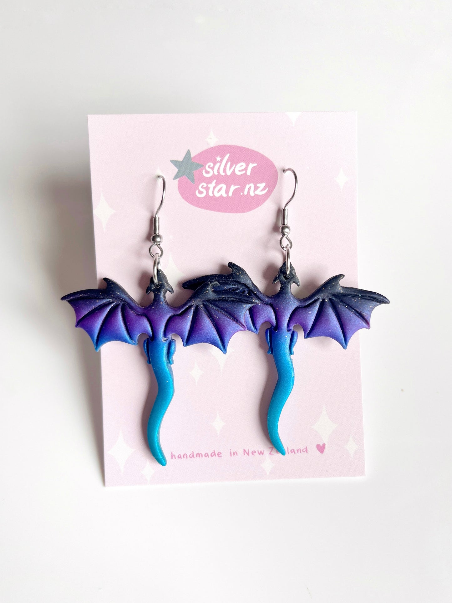 The Blue Dragon Polymer Clay Earrings feature funky dragon shapes with purple wings and blue tails, displayed on a card saying silverstar.nz and handmade in New Zealand with a heart symbol.
