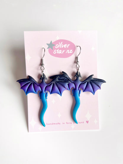 The Blue Dragon Polymer Clay Earrings feature funky dragon shapes with purple wings and blue tails, displayed on a card saying silverstar.nz and handmade in New Zealand with a heart symbol.
