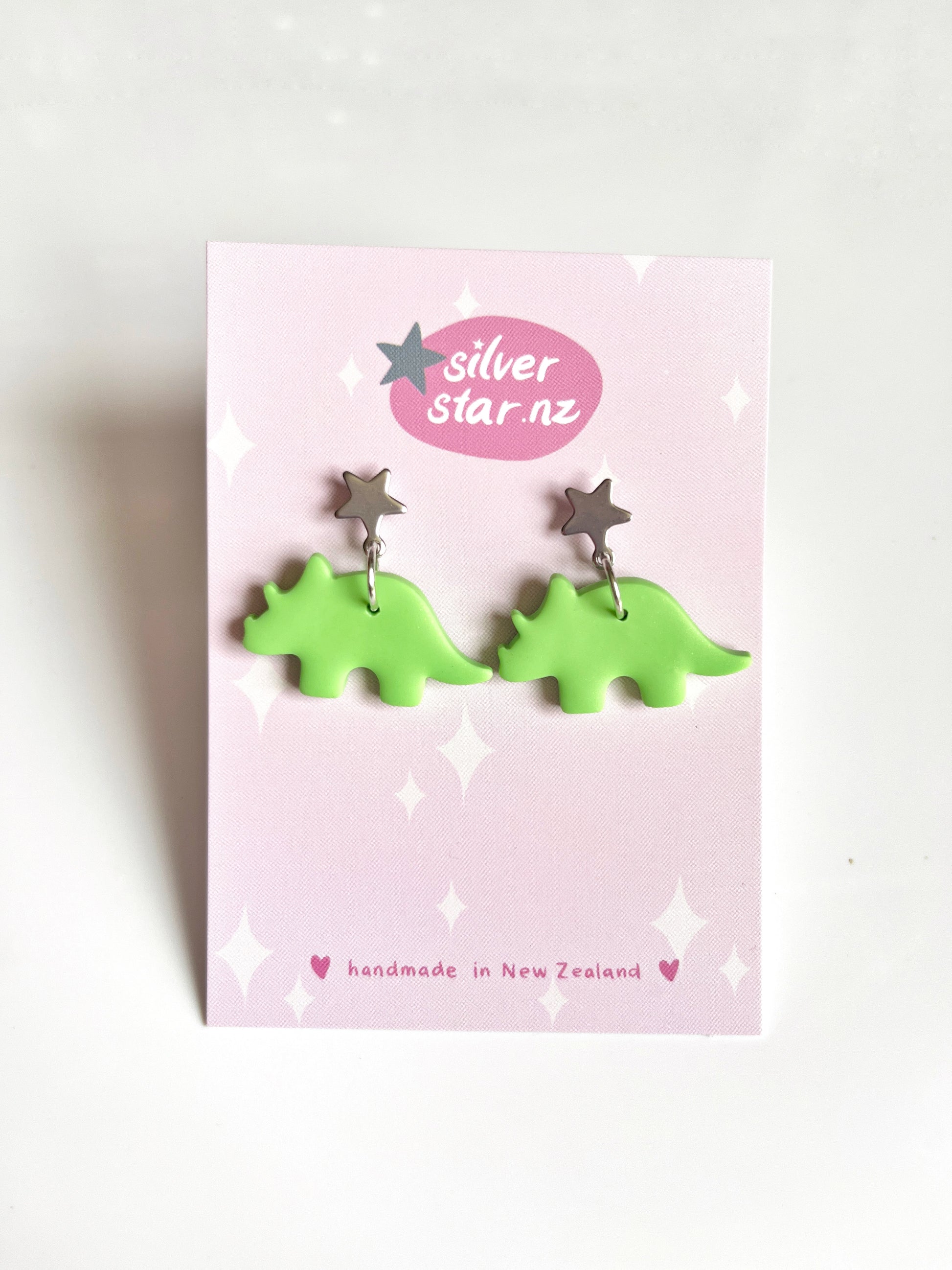 The pink card features Dino Polymer Clay Earrings with green dinosaur shapes and star-shaped studs. Handmade charm oozes from these unique earrings. The silverstar.nz logo is present, with a note: handmade in New Zealand. Its set against a plain white background.