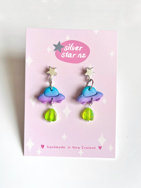 Handmade UFO Polymer Clay Earrings on a pink card feature blue and purple clouds, bright yellow-green raindrops, and a silver star. Crafted with stainless steel hooks, the card displays silverstar.nz and handmade in New Zealand.