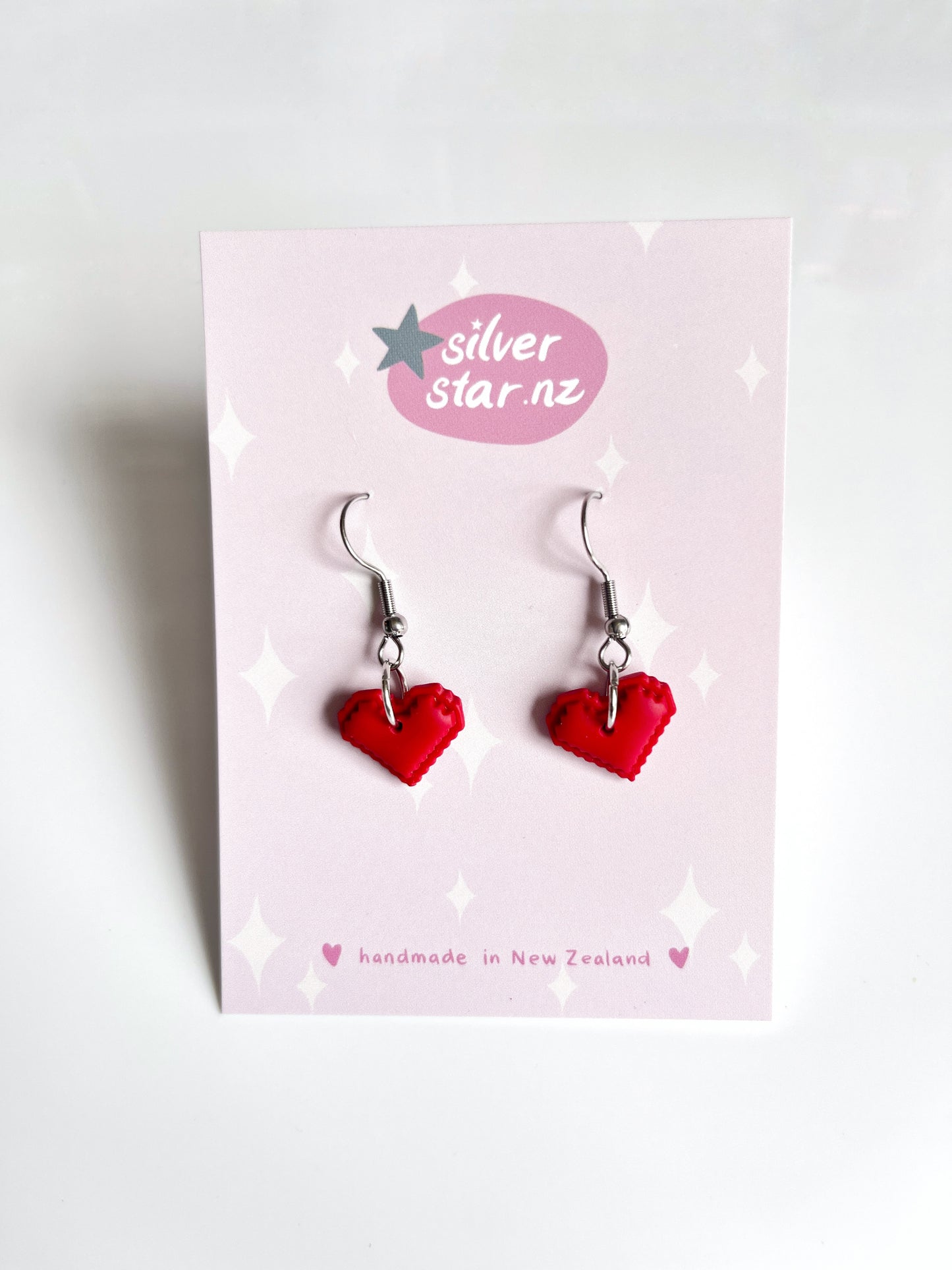 The Pixel Heart Polymer Clay Earrings, vibrant red and uniquely heart-shaped, are displayed on a pink card with a silver star logo and silverstar.nz. The card highlights they are handmade in New Zealand.