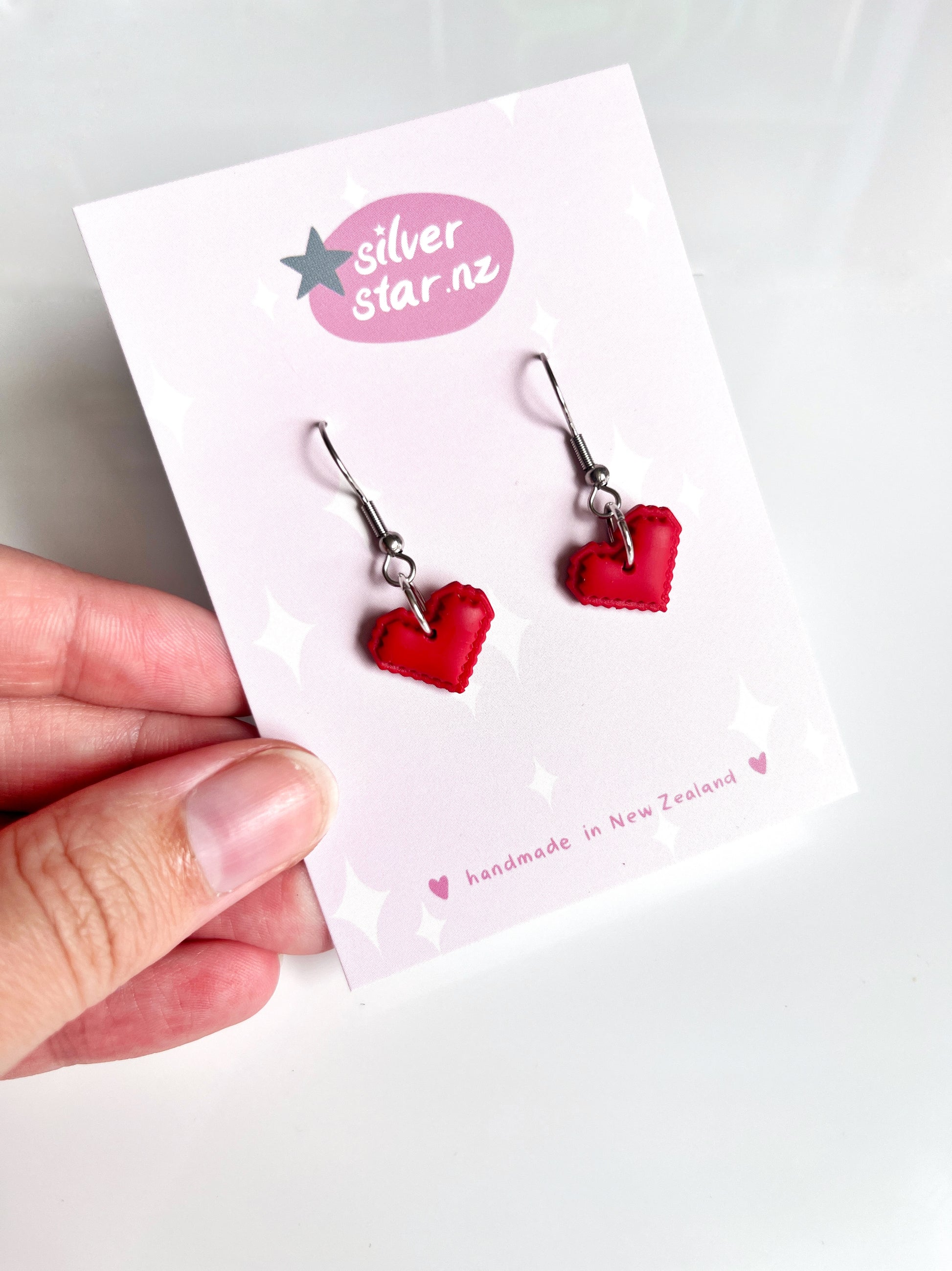 A hand displays a card with silverstar.nz and red Pixel Heart Polymer Clay Earrings, highlighting their handmade quality in New Zealand. These uniquely designed earrings embody artistry and love.