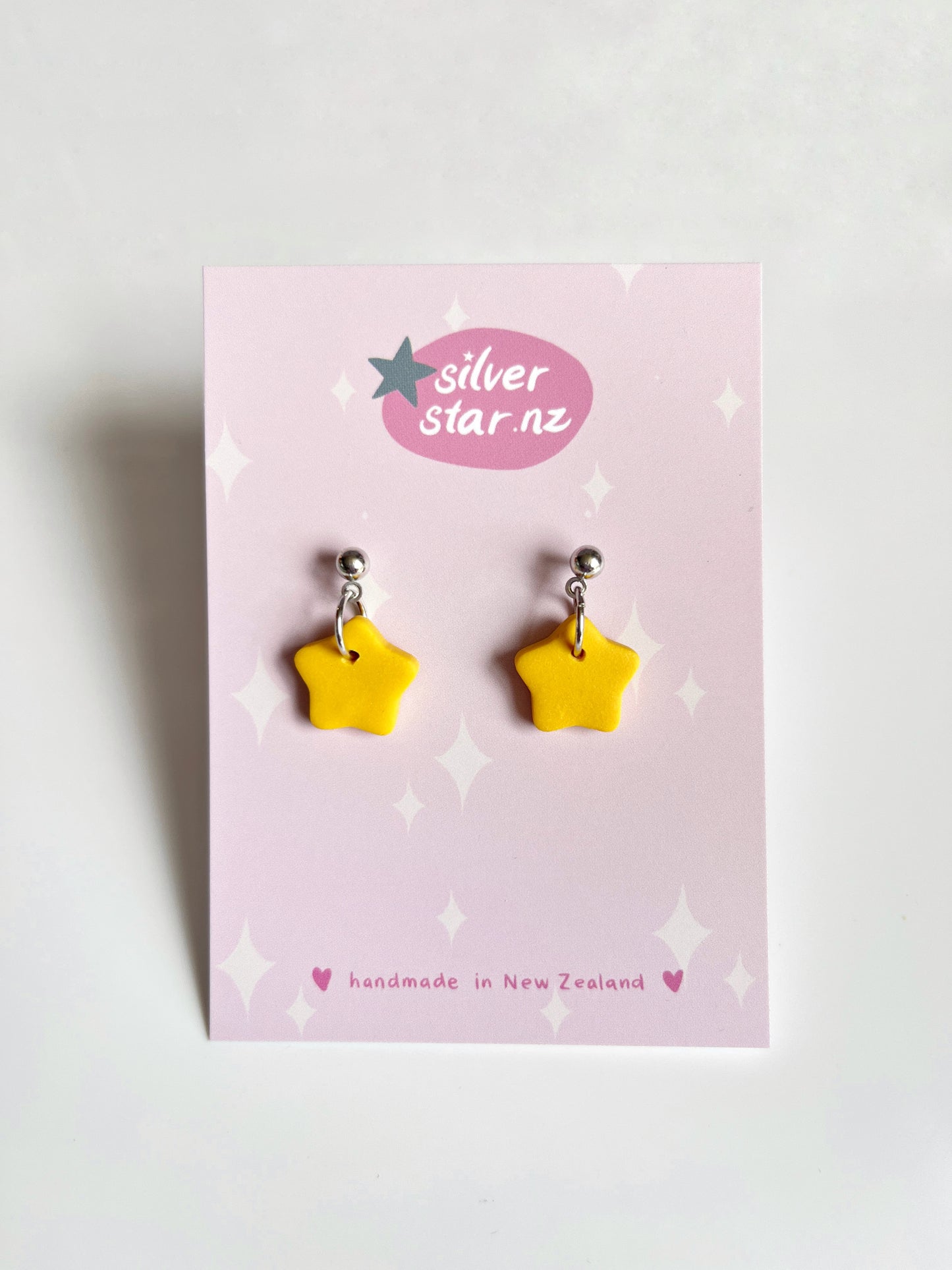 Yellow Star Polymer Clay Earrings from silverstar.nz are presented on a pink star-patterned card, showcasing their handmade New Zealand craftsmanship. Each vibrant earring features stainless steel hooks for durability.