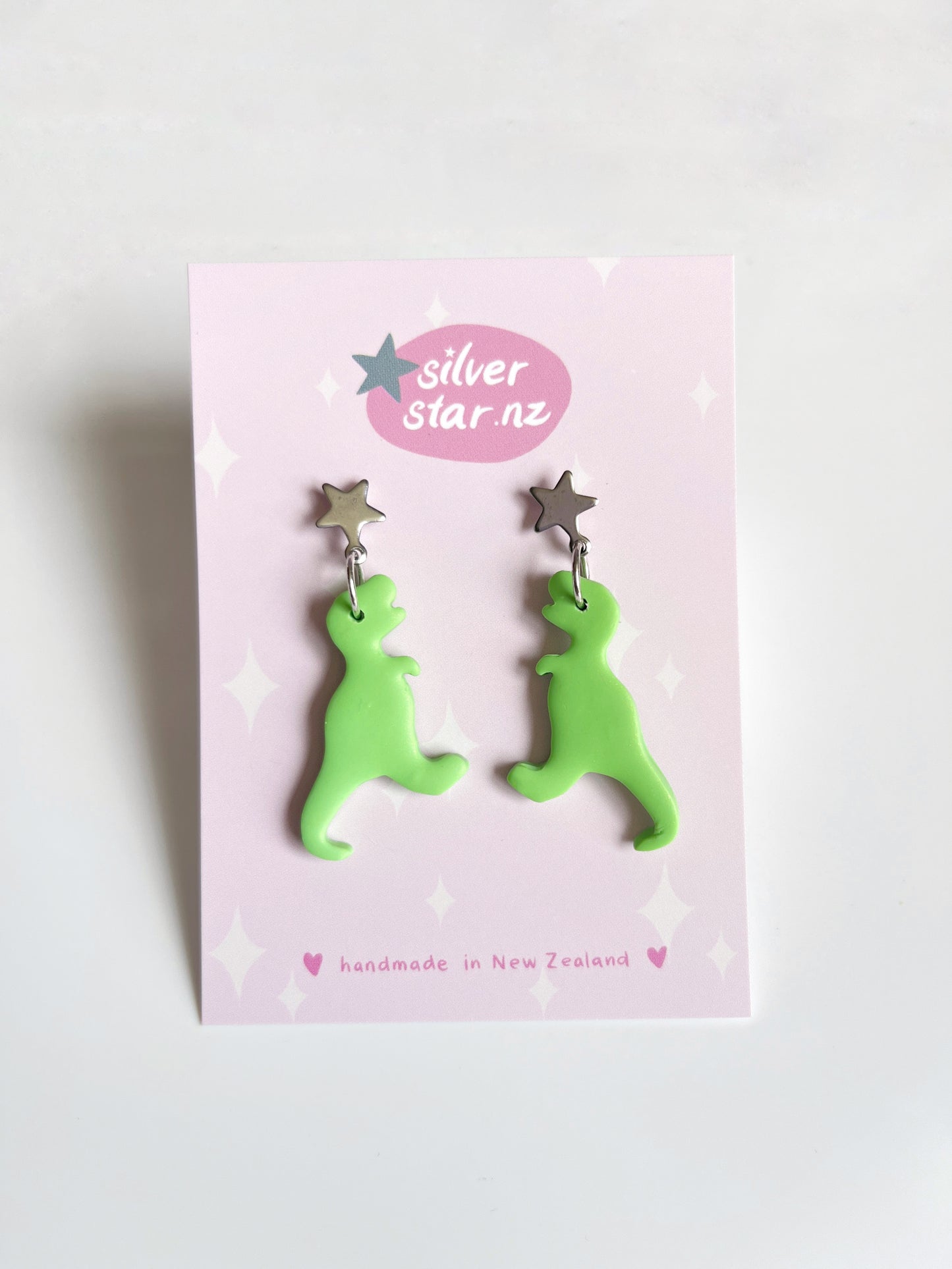 The Dino Polymer Clay Earrings by silverstar.nz are unique green dinosaur designs with star-shaped studs, presented on a pink card featuring the logo and handmade in New Zealand, showcasing exquisite craftsmanship.