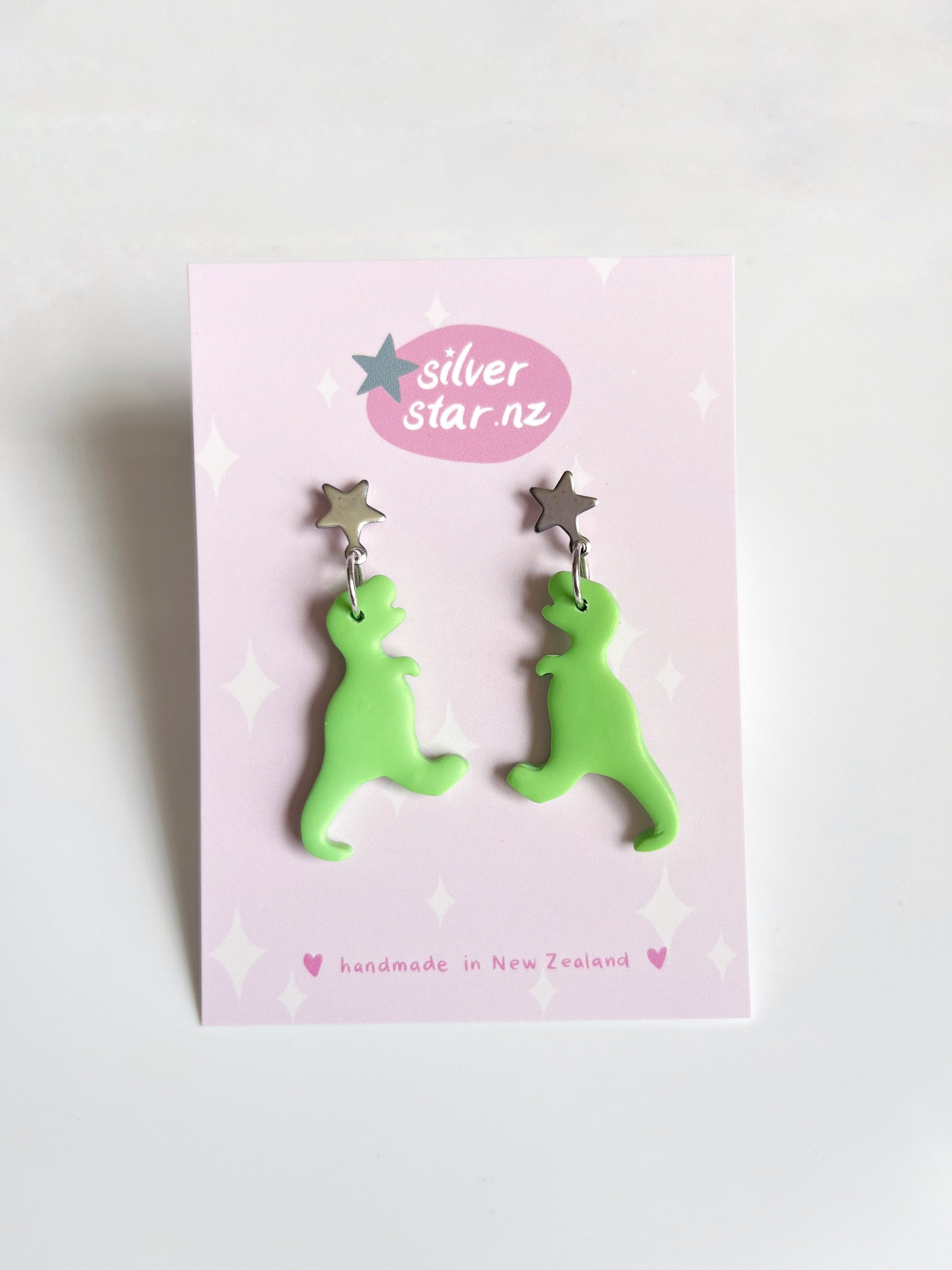 The Dino Polymer Clay Earrings by silverstar.nz are unique green dinosaur designs with star-shaped studs, presented on a pink card featuring the logo and handmade in New Zealand, showcasing exquisite craftsmanship.
