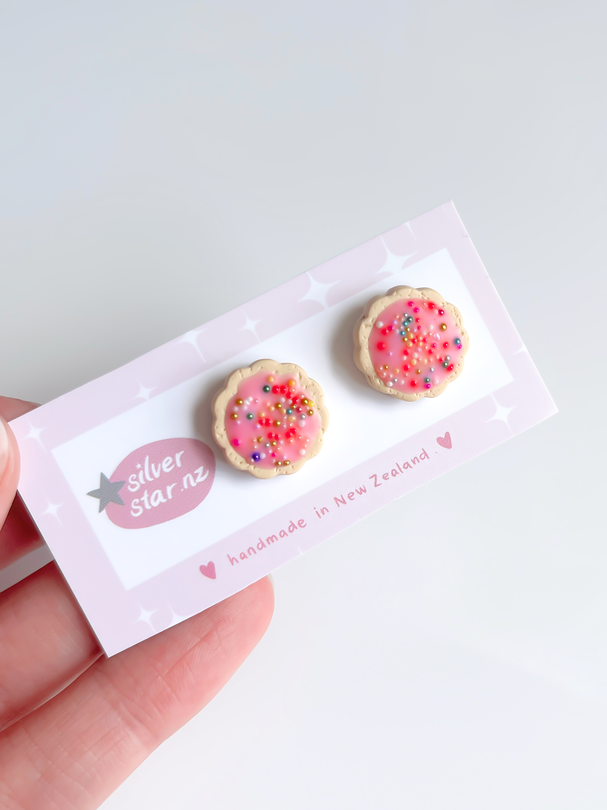 A hand holds the Hundreds and Thousands Biscuit Stud Earrings, unique handmade polymer clay earrings resembling pink frosted cookies with sprinkles. They are attached to a card from silverstar.nz reading handmade in New Zealand, highlighting creative craftsmanship.
