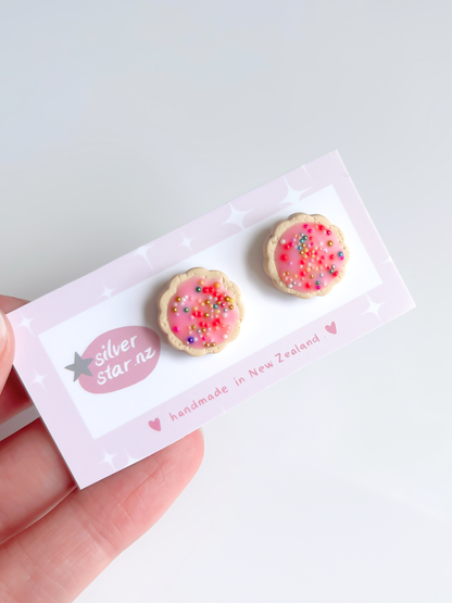 A hand holds the Hundreds and Thousands Biscuit Stud Earrings, unique handmade polymer clay earrings resembling pink frosted cookies with sprinkles. They are attached to a card from silverstar.nz reading handmade in New Zealand, highlighting creative craftsmanship.