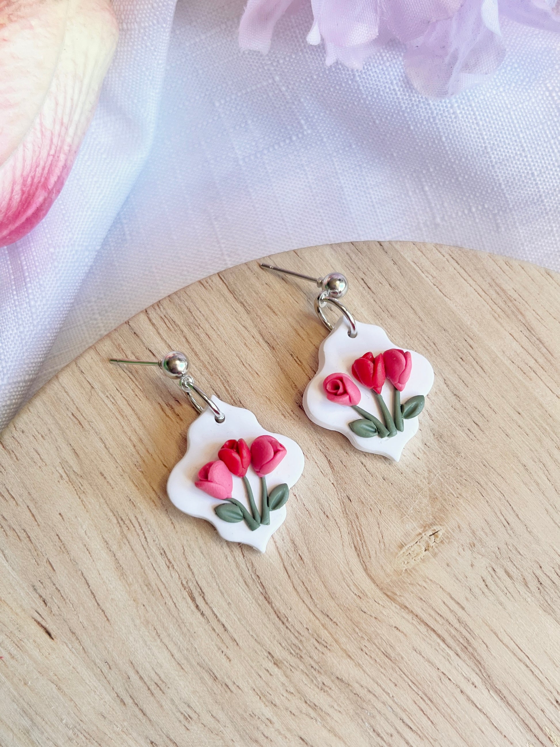 The Tulip Floral Polymer Clay Earrings from silverstar.nz, with a white base and red-pink floral designs, rest on wood. Handmade with durable stainless steel hooks, they are complemented by soft pink sheer fabric nearby, adding elegance to the scene.