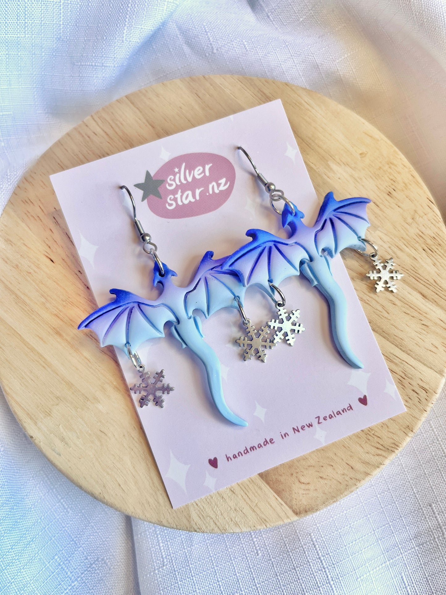 The Winter Snow Dragon Polymer Clay Earrings by silverstar.nz feature intricate blue dragon designs and dangling silver snowflakes, crafted with polymer clay and set on stainless steel hooks. Displayed on wood, the card reads silver star.nz and handmade in New Zealand.
