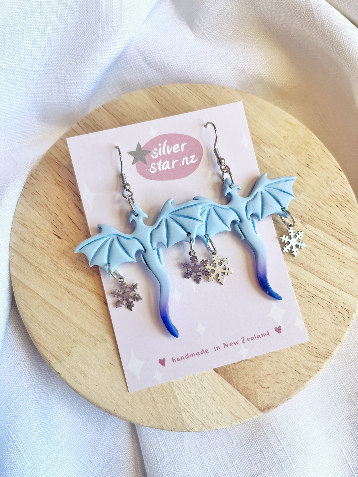 Displayed on a circular wooden surface, the Winter Snow Dragon Polymer Clay Earrings from silverstar.nz enchant with their light blue dragon shape and star charms. Crafted with stainless steel hooks, these handmade New Zealand pieces come on a branded card against an elegant white cloth backdrop.