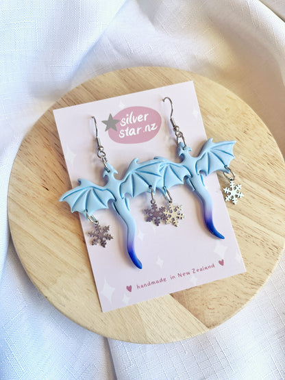 Displayed on a circular wooden surface, the Winter Snow Dragon Polymer Clay Earrings from silverstar.nz enchant with their light blue dragon shape and star charms. Crafted with stainless steel hooks, these handmade New Zealand pieces come on a branded card against an elegant white cloth backdrop.