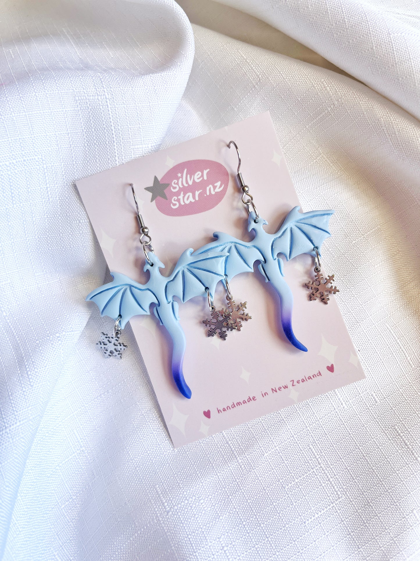 The Winter Snow Dragon Polymer Clay Earrings from silverstar.nz feature handmade blue dragon shapes with intricate details, accented by silver snowflake charms on stainless steel hooks, elegantly displayed on a white textured fabric background.
