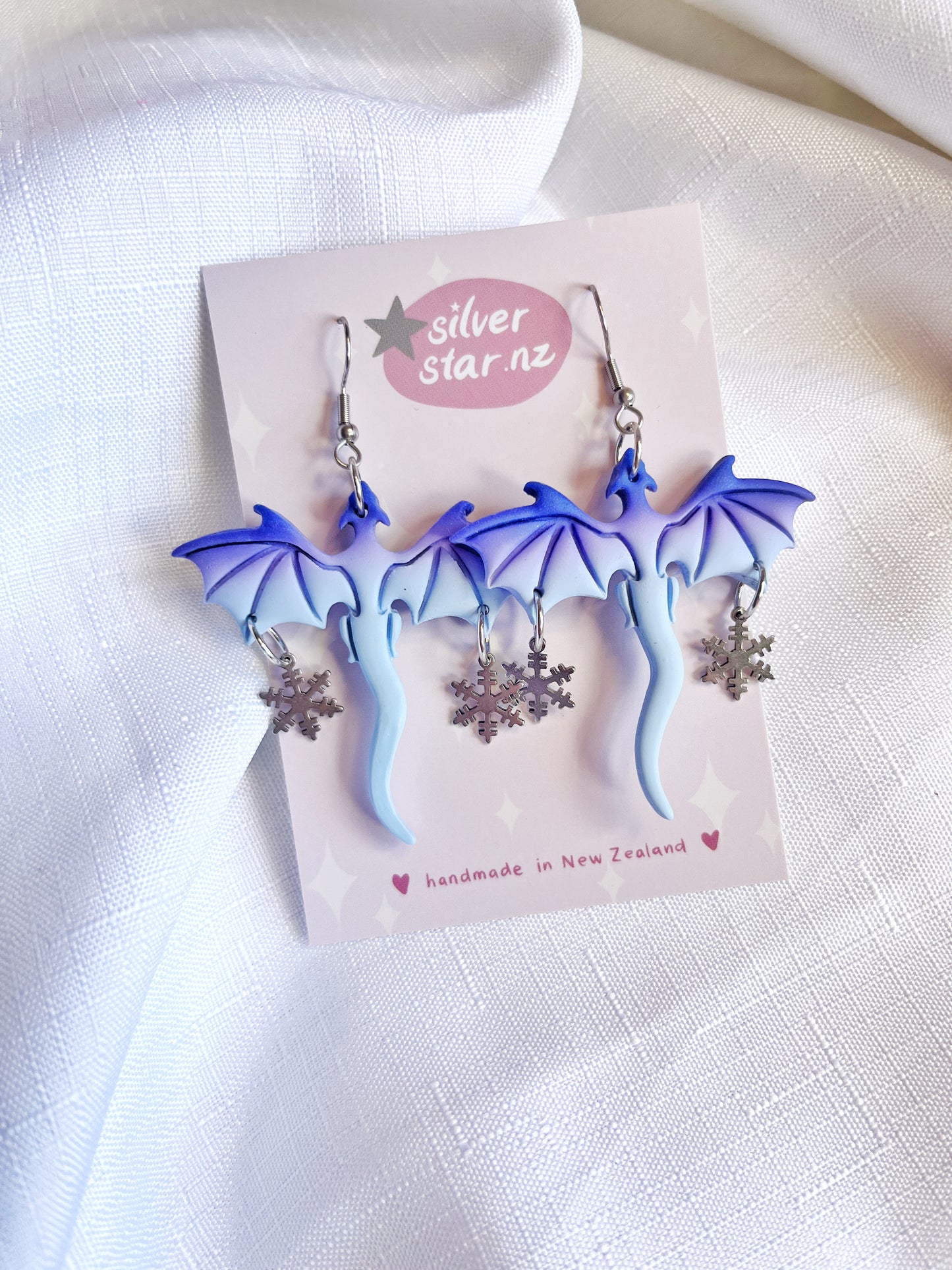 Winter Snow Dragon Polymer Clay Earrings from silverstar.nz showcase blue dragon charms with dangling silver snowflakes on a silverstar.nz card, set against a white fabric backdrop. These handmade New Zealand earrings feature durable stainless steel hooks.