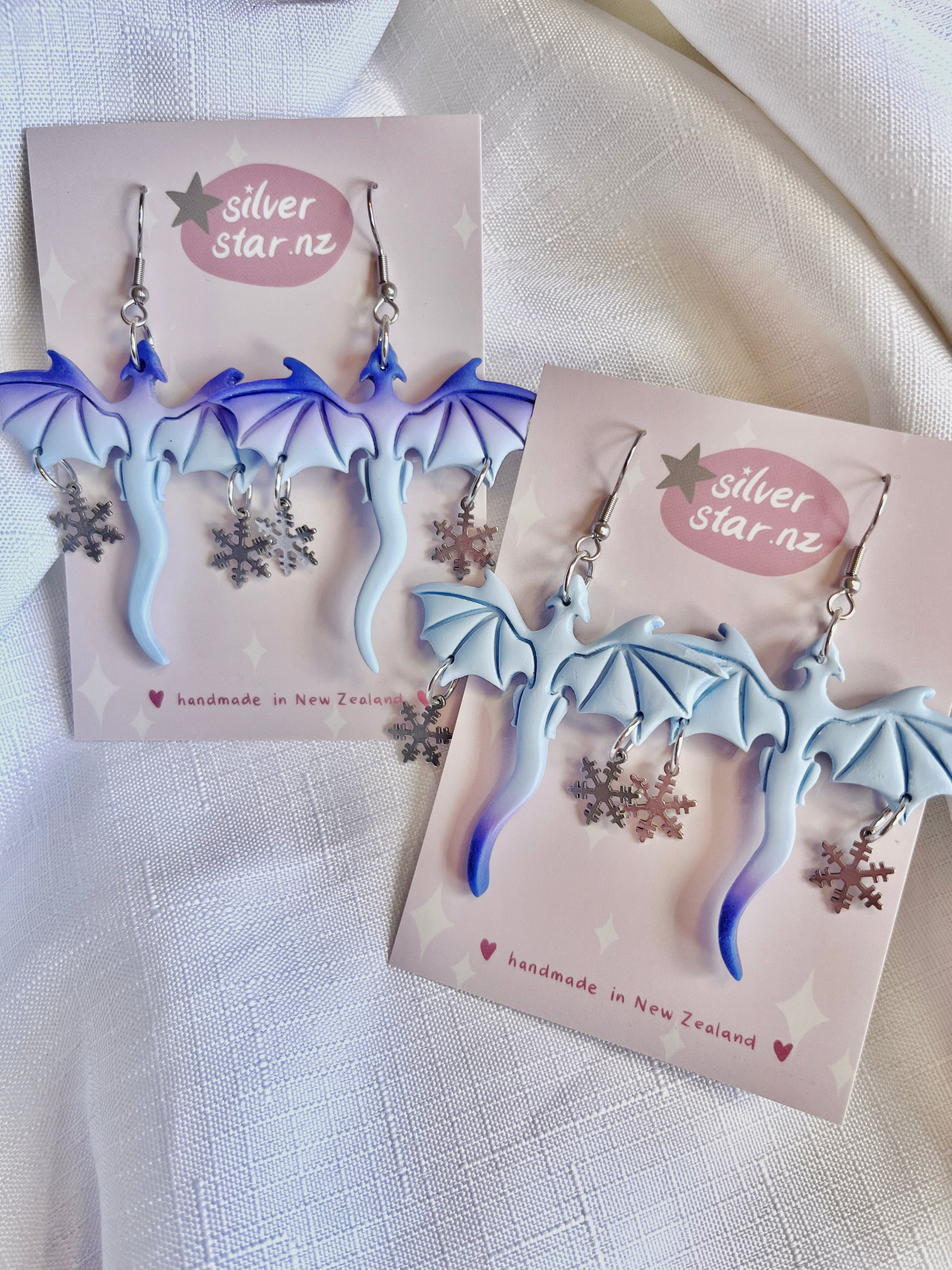 The Winter Snow Dragon Polymer Clay Earrings from silverstar.nz are handmade in New Zealand, featuring blue and purple gradient dragon designs with snowflakes dangling, and stainless steel hooks. They are displayed on silverstar.nz labeled cards.