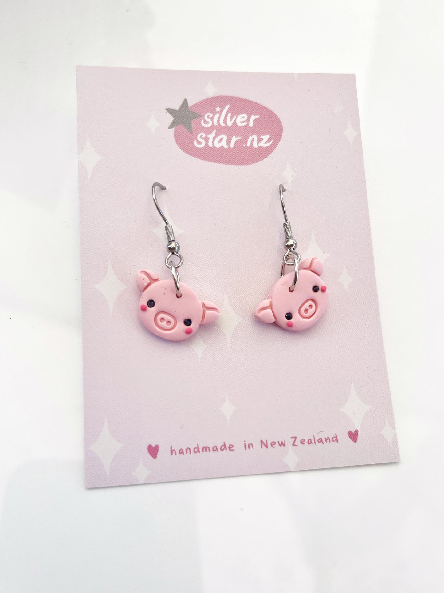 A pair of cute pink pig-shaped polymer clay earrings with stainless steel hooks is showcased on a card decorated with a whimsical pink and white star pattern, featuring silverstar.nz and handmade in New Zealand.