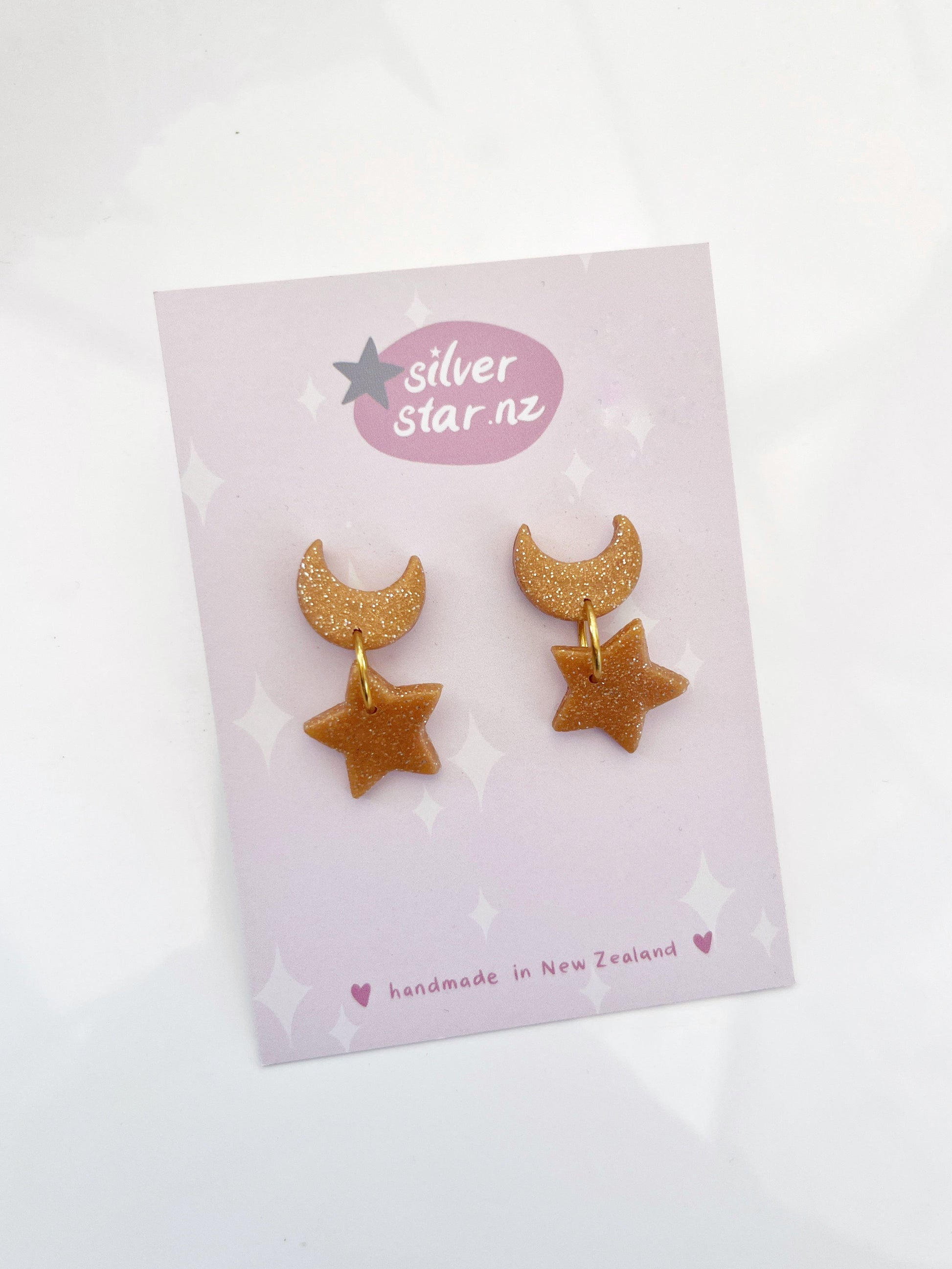 The Star + Moon Polymer Clay Earrings, with a glittery crescent moon and star design, hang from stainless steel hooks. Displayed on a card, they highlight the brand silverstar.nz and proudly note their handmade origin in New Zealand.
