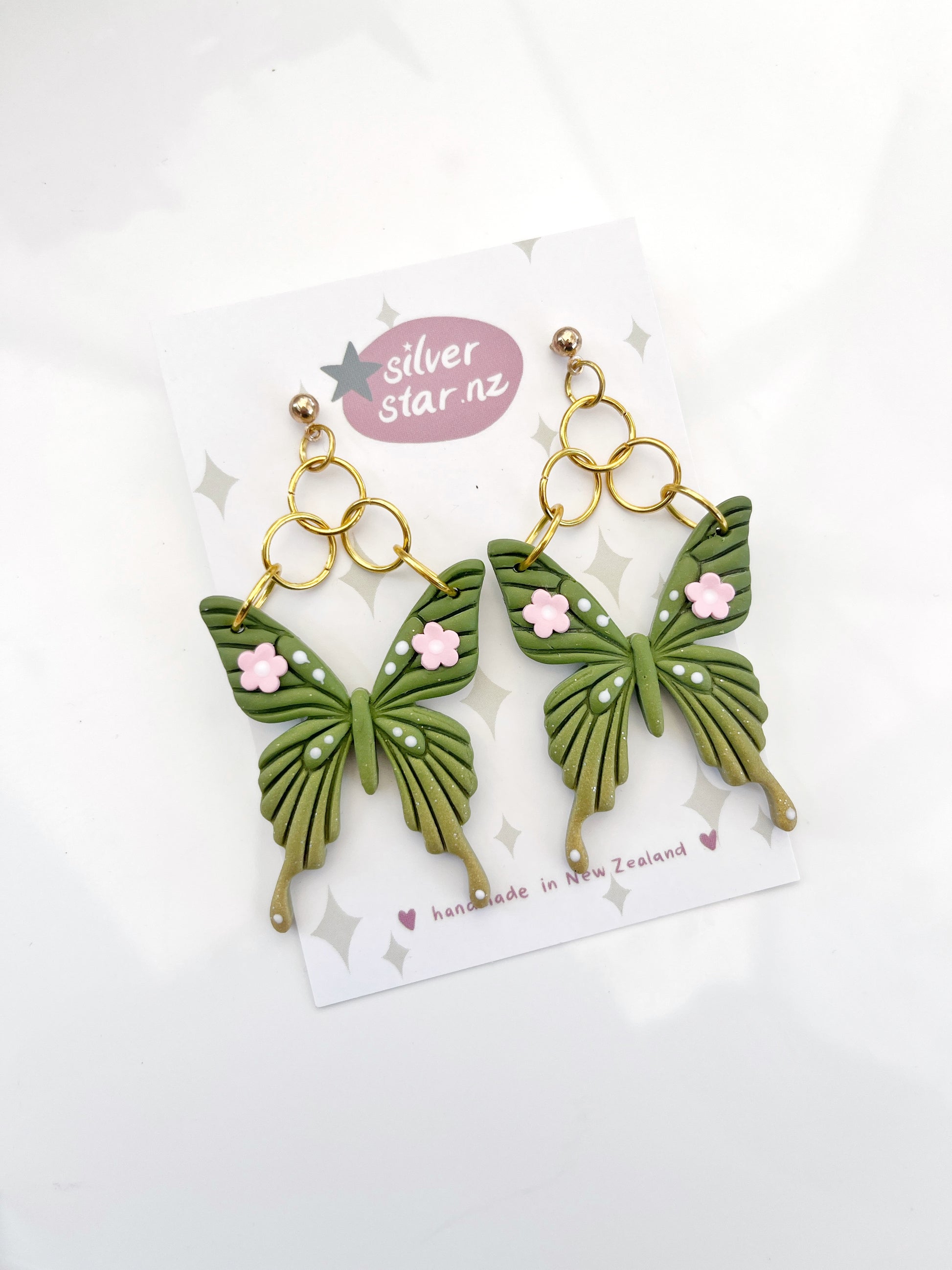 The Green Fae Butterfly Polymer Clay Earrings by silverstar.nz feature a unique design with green butterflies and pink floral accents. Displayed on a branded card, they showcase intricate wing patterns, gold-colored hooks, and are lovingly handmade in New Zealand.