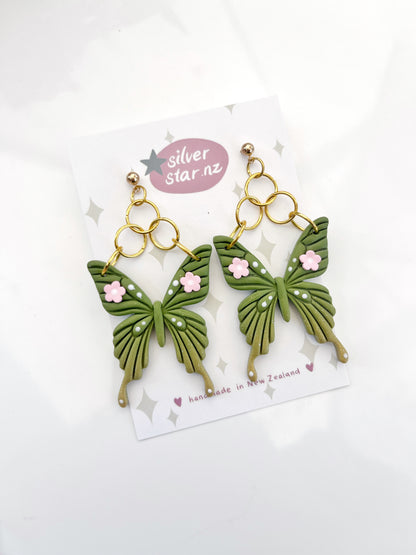 The Green Fae Butterfly Polymer Clay Earrings by silverstar.nz feature a unique design with green butterflies and pink floral accents. Displayed on a branded card, they showcase intricate wing patterns, gold-colored hooks, and are lovingly handmade in New Zealand.