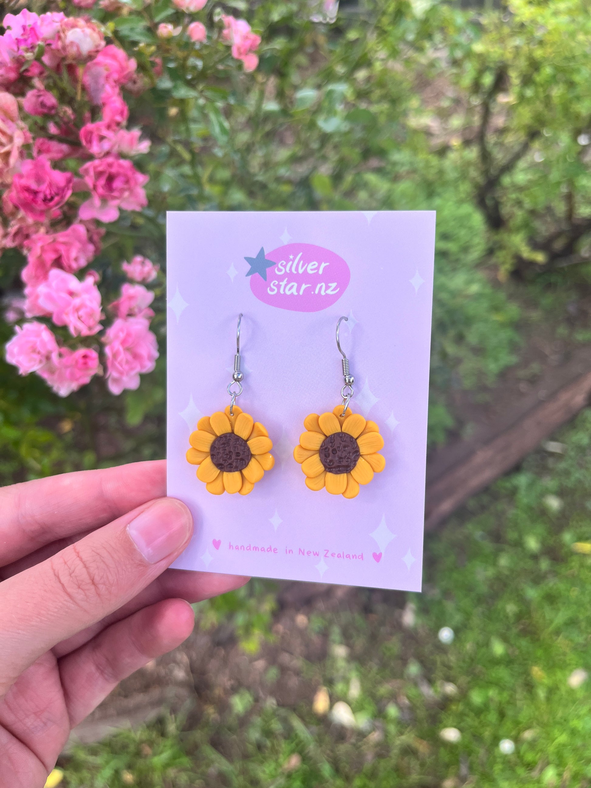 A hand displays sunflower-shaped Sunflower Polymer Clay Earrings on a card with decorative stars, surrounded by vibrant pink flowers and green foliage. The card reads silverstar.nz, indicating exquisite handmade earrings crafted in New Zealand.