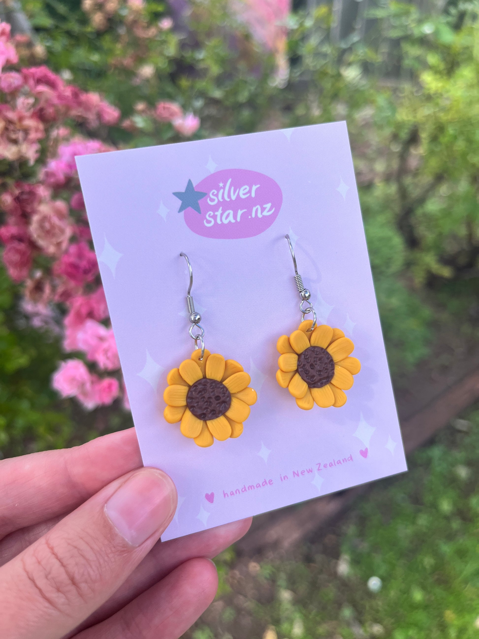 A hand showcases sunflower polymer clay earrings on a silverstar.nz branded card. The background features blurred pink flowers and greenery and the card states handmade in New Zealand.