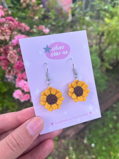 A hand showcases sunflower polymer clay earrings on a silverstar.nz branded card. The background features blurred pink flowers and greenery and the card states handmade in New Zealand.