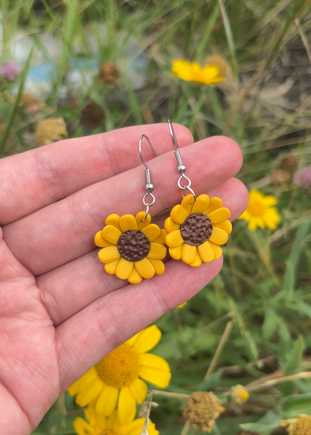 A hand holds a pair of Sunflower Polymer Clay Earrings by silverstar.nz, crafted with vibrant yellow petals and rich brown centers. Green plants and scattered yellow flowers provide an enchanting background.